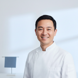 Portrait of a smiling male chef, David Wu, wearing a white chef's jacket against a clean, light blue and white background.